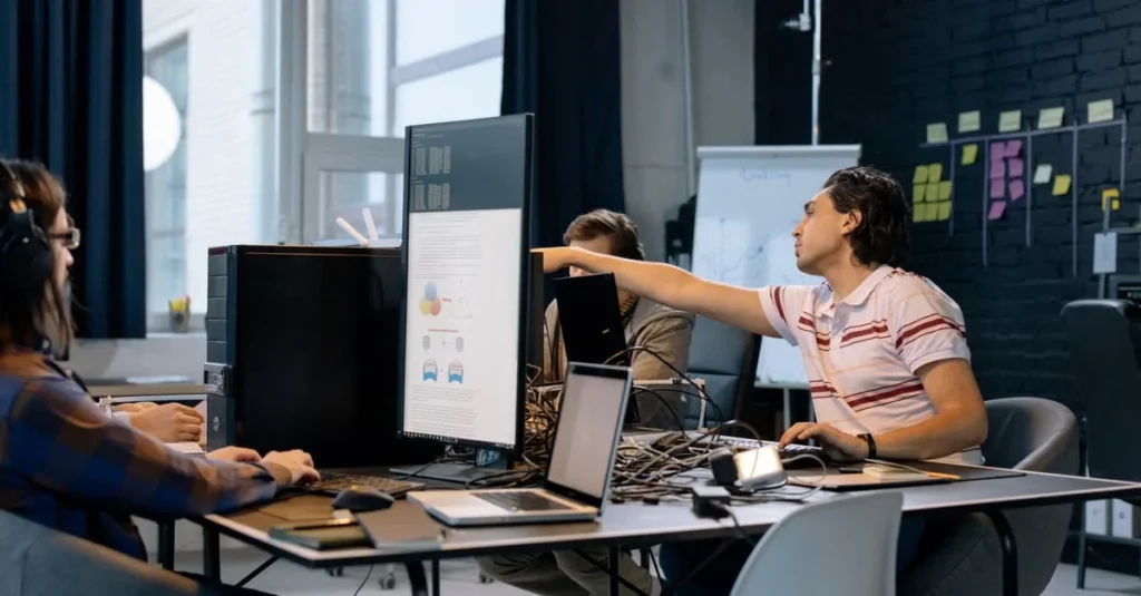 Team collaborating at desk with computer monitors and sticky notes on dark wall in modern office space.