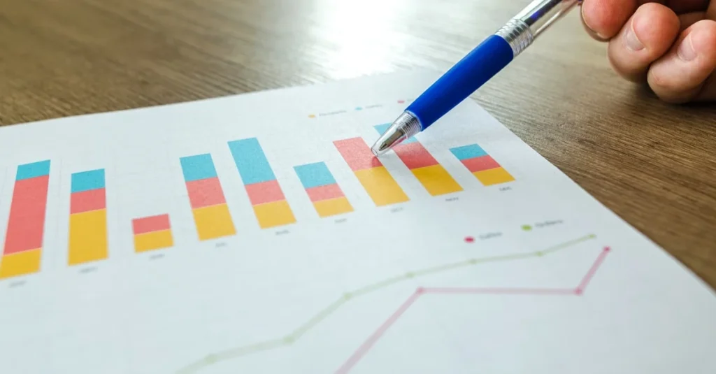 Close-up of a hand using a blue pen to point at colorful bar and line charts on a printed report, suggesting business data analysis.