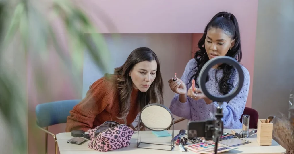 Two women focus on a makeup tutorial at a table, surrounded by cosmetics, mirrors, and a ring light in a warm, creative setting.