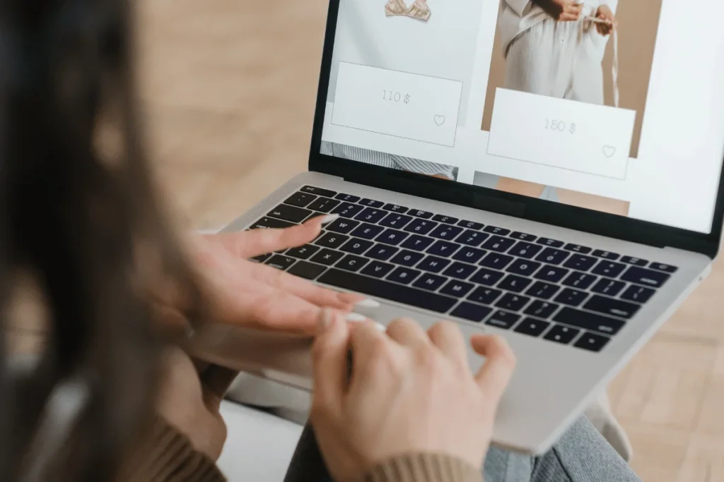 Person browsing an online fashion store on a laptop, viewing clothing photos and prices while typing; warm, modern shopping scene.