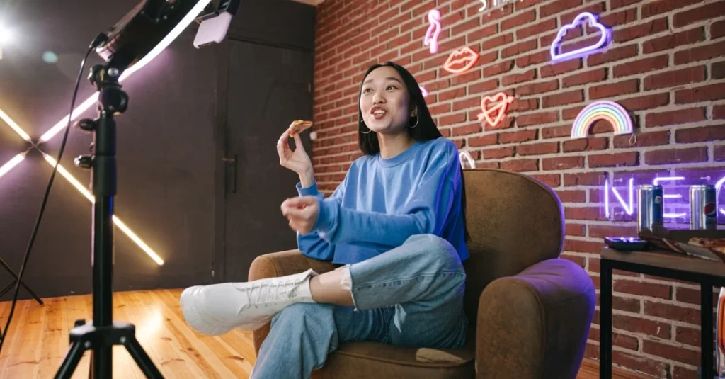 Woman in a blue sweater sits in an armchair speaking to a camera in a neon-lit brick studio, creating a relaxed, upbeat mood.