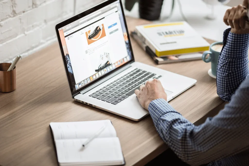 Person in a checkered shirt works on a laptop displaying website design at a wooden desk with a notebook, coffee, and design books.