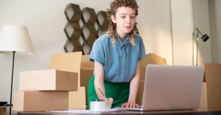 Young woman managing boxes and using affiliate tracking software on laptop for ecommerce business operations.